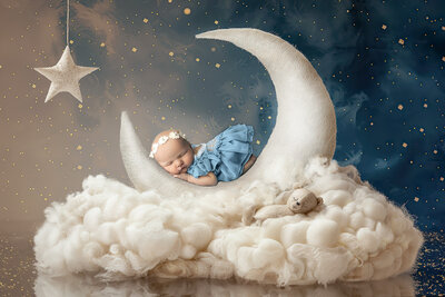 Newborn baby girl peacefully sleeping on a white crescent moon prop surrounded by fluffy clouds and a starry night sky, wearing a blue dress and floral headband. Captured by a Dallas Newborn Photographer.