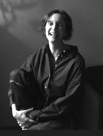 Black and white photo of woman with short hair smiling vibrantly. She is sitting in a chair in a button down shirt, holding one leg up.