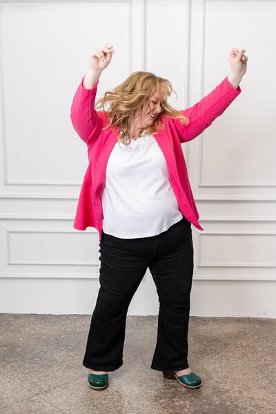 Woman in a bright pink blazer dances with arms raised during a personal branding photoshoot in a modern white studio. This energetic image captures her fun, confident personality—perfect for showcasing an approachable and spirited brand.