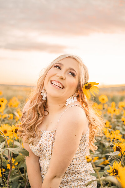 blonde senior girl with a sunflower behind her ear laughing in a patch of flowers at sunset