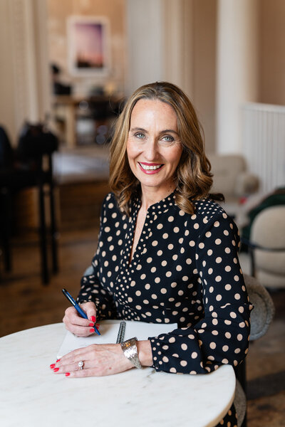 woman in black and beige polka dot dress, sitting at a desk and writing to her notebook, looking at the camer