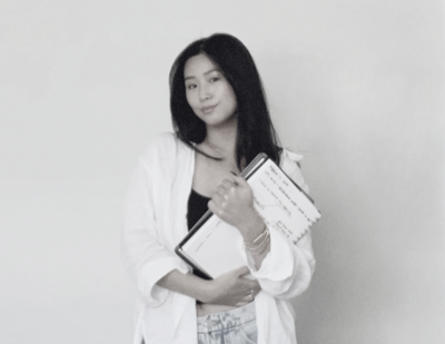 Shiho, graphic designer at Interiors by CV, holding design materials and wearing a light jacket in a studio portrait.