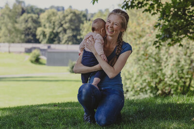 En kvinne står på et kne i en park og holder babyen sin over skulderen mens hun ler. Kvinnen har på blå dongeribukse og blå t-skjorte. Flettet hår.
