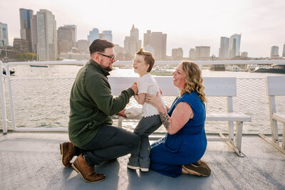 Two parents are kneeling on the floor of the Charlestown ferry, holding their 10 year old daughter, who is laughing. The Boston skyline is glowing behind them.