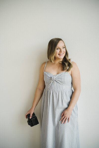 A photo of Krista Marie, standing in front of a while wall with her camera in her hand, smiling and looking away from the camera