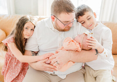 Dad holding newborn on a couch while sister looks at the camera and brother feels baby's head