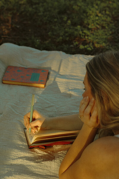 A woman is facing away from the camera and you can see her resting her head on her hand as she is laying down to write in her journal. A book rests on the blanket she is laying on in the background