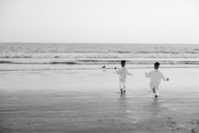 Black and white photo of two young boys in white outfits running toward the ocean on a sandy beach.