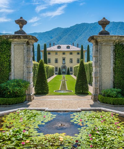 A grand villa nestled at the end of a manicured lawn, bordered by tall hedges. A pathway leads to the entrance, and a decorative pond with water lilies and a fountain is in the foreground. Mountains rise in the background under a clear blue sky.