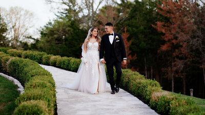 A bride and groom walk hand in hand down a stone path, smiling at each other, surrounded by lush greenery.