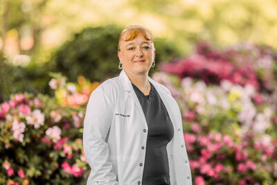 woman wearing a white doctor's coat standing in front of azalea bushes while smiling