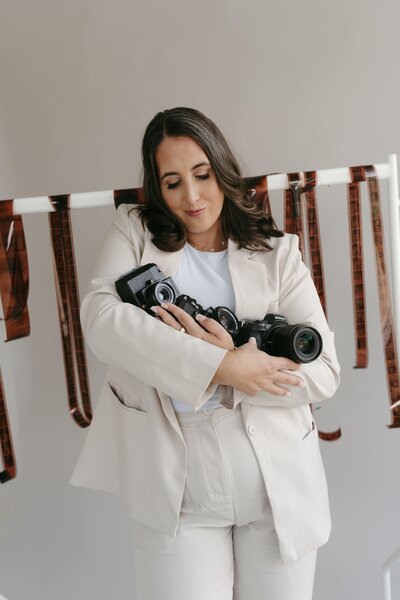 Professional wedding photographer in a cream suit holding film and digital cameras, standing in front of a backdrop decorated with hanging film strips.