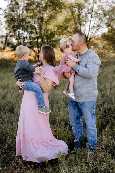 Mom dancing with her son in a green grass field, dad holding his one year old daughter kissing her on the forhead.
