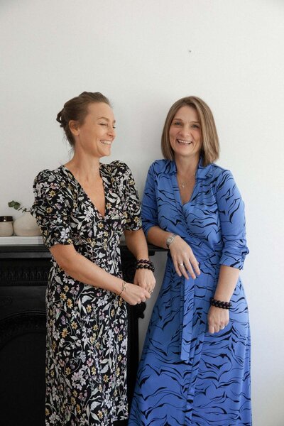 Naturopath Lauren Glucina and spiritual healer Sharon Emery are standing laughing by a modern black fireplace.