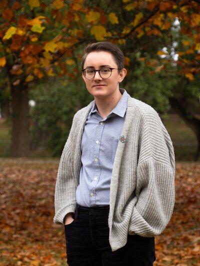 professional headshot for a young queer person, they wear a button down and cardigan with a pride pin and stand against a backdrop of autumn leaves at the Christchurch Botanic Gardens in New Zealand.