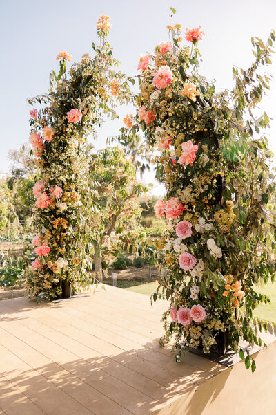 A gorgeous oversized wedding ceremony arch is flush with tropical florals of pinks, whites and yellows for a Flora Farms Wedding in Los Cabos Mexico