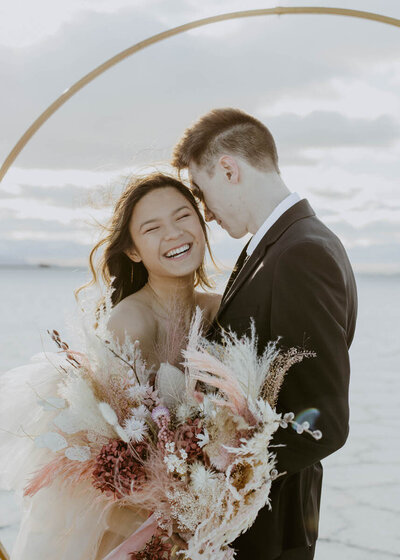 Bride and groom holding each other laughing while holding pink and tan bouquet under a gold ceremony arch at the Salt Flats in Utah.