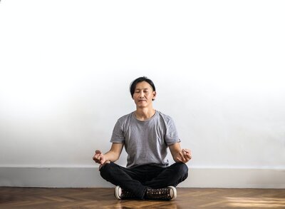 A man sits on a wooden floor, legs crossed and eyes closed. He rests his hands on his knees and has a soft, serene expression on his face.