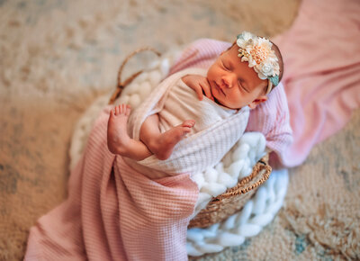 Newborn baby has hand on chin while wrapped in muted blue swaddle and hat with ears laying on flowing blanket in a basket peacefully sleeping.