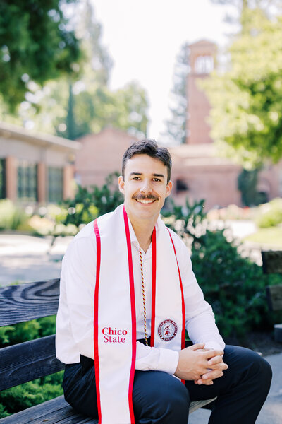 Chico State senior graduate wearing cap and gown, celebrating graduation on campus with Chico State University buildings and greenery in the background.