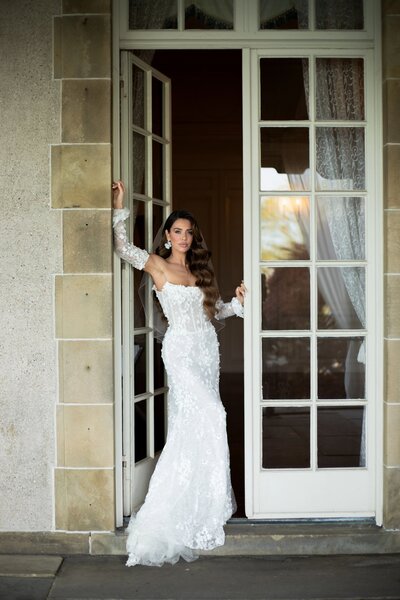 Rhode Island Wedding Photographer | Kelsey Sheehan Photography Timeless Rhode Island Weddings | A woman in an elegant, lace off-shoulder white gown stands confidently in a doorway. The setting is serene, with soft, natural light enhancing a romantic tone.