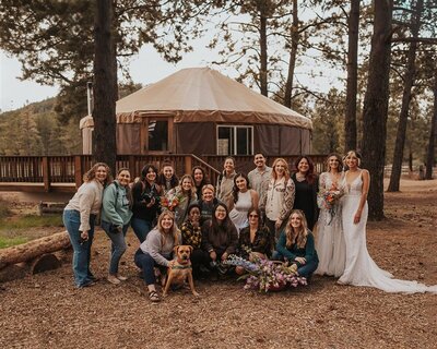 A group of photographers, along with two individuals in wedding outfits, pose in front of a yurt in a wooded area. The group is smiling and holding bouquets, with a dog sitting happily in the front.