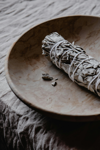 A bundle of sage sits on an organic ceramic dish atop a table with a linen crinkled tablecloth.