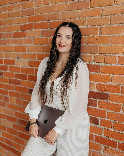 Taysha smiling at the camera holding her closed laptop leaning against a red brick wall.