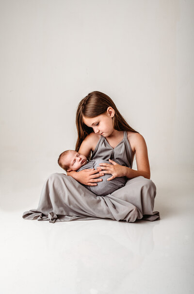 Family photographers Maryland captures newborn pictures with older sibling holding baby while they both wear grey dresses for an in studio newborn photo shoot
