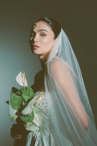 A bride in a vintage-inspired lace gown and sheer veil holds a bouquet of white and green anthuriums, softly lit with dramatic shadows, evoking a timeless, ethereal elegance