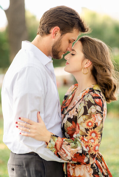 Portrait of a man and woman in a white shirt and floral dress leaning into each other outdoors with greenery in the back.