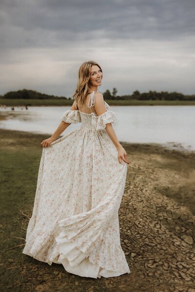 A woman in a long, flowing floral dress stands by a lake on a cloudy day, looking back over her shoulder and smiling—captured beautifully by a family photographer in Macon GA.