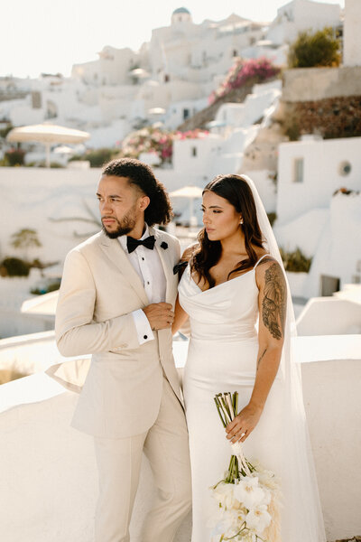 Bride and groom standing together in Santorini with the iconic whitewashed cliffside village and bougainvillea in the background.
