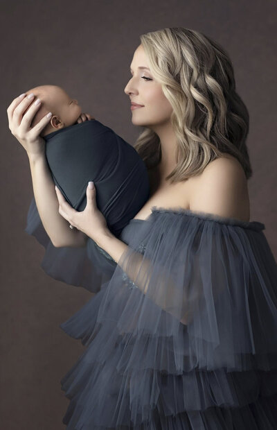 8 day newborn girl wrapped in a forest green blanket with mother holding baby and wearing a dusty-colored blue gown.