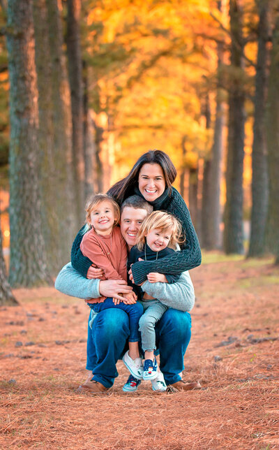Outdoor family photos in the woods with the father kneeling down holding his children close in the mother leaning over the father shoulders and hugging them all while smiling captured by Family photographer Hampton Roads va