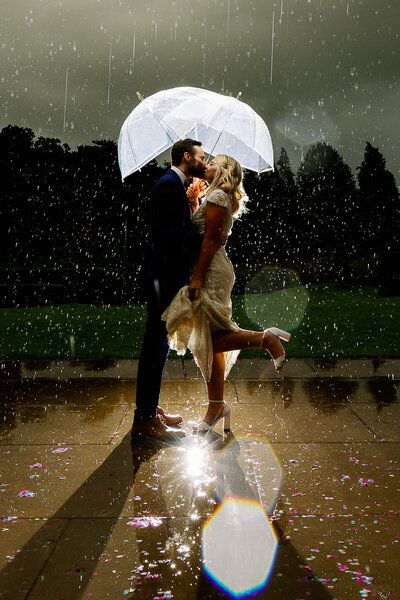 A bride and groom kissing in the rain under umbrellas on the grounds of Utopia at the Broughton Hall Sanctuary