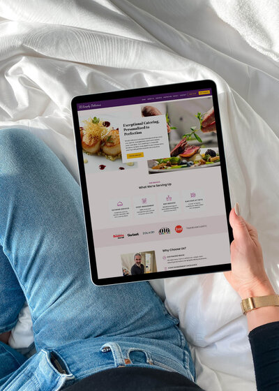 A hand holds an iPad displaying the Simply Delicious Catering website.