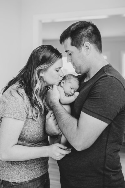 Family snuggles and kisses newborn baby girl in black and white photo during in-home newborn session