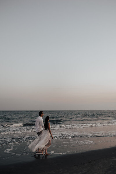 Charleston beach engagement photo. Couple walking at the beach right at the water. DRess flowing back. Captured right at sunset