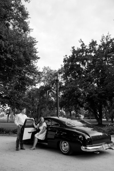 Black and white photo of a couple posing with a vintage car on their wedding day.