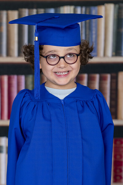 Toddler boy wearing white buttondown, suspenders, and teal bowtie smiles sweetly for the camera and his school photos.