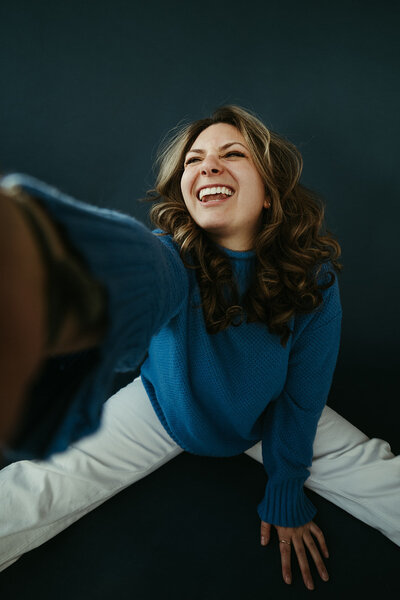 Sara Dobbins sits on blue backdrop wearing a blue sweater. She smiles big and reaches right hand towards camera