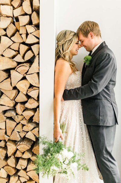 Bride and groom touch noses and embrace against a white wall near a stack of wooden logs