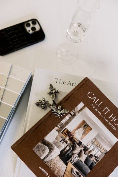 A stack of books titled "Call It Home" and "The Art" with a metallic bee decoration on top. Nearby, there's a smartphone with a black case, a glass of water, and paint swatches on a white surface.