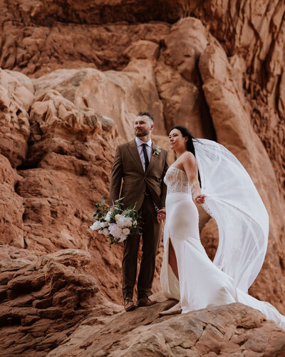 bride and groom standing on large red rock formation in the desert