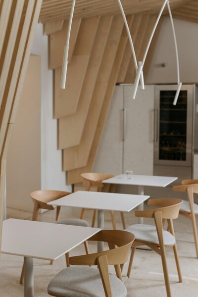 A welcoming café interior featuring three sleek white tables, each accompanied by wooden chairs with white leather-cushioned seats. The thoughtfully arranged seating provides a cozy yet modern atmosphere, perfect for customers to enjoy freshly baked goods and specialty coffee.