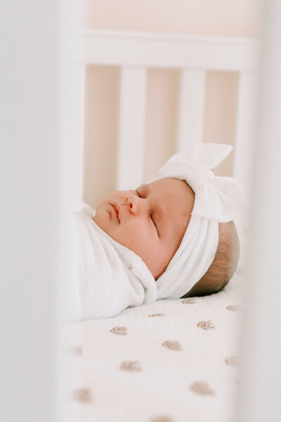 A newborn baby taking a nap in her crib while swaddled in a blanket with a bow on her head