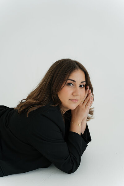 Woman laying on her stomach, resting her head on hands, wearing black blazer in bright studio.