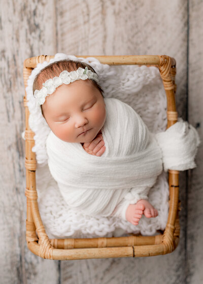baby girl sleeping in a basket wearing a white swaddle and a white flower headband