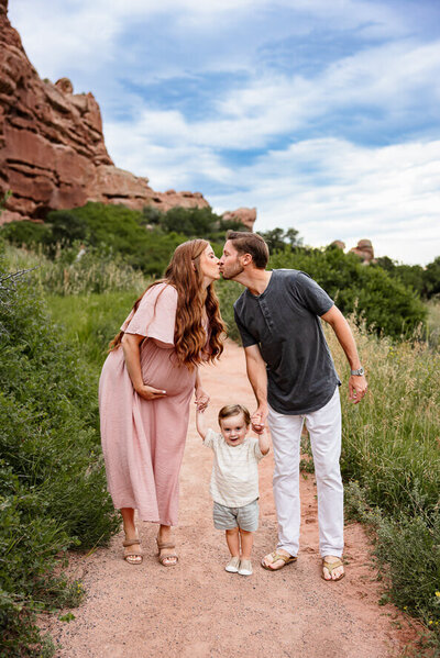 A pregnant woman in a pink dresses leans over to kiss her husband, while holding hands with her toddler son in front of red rocks.